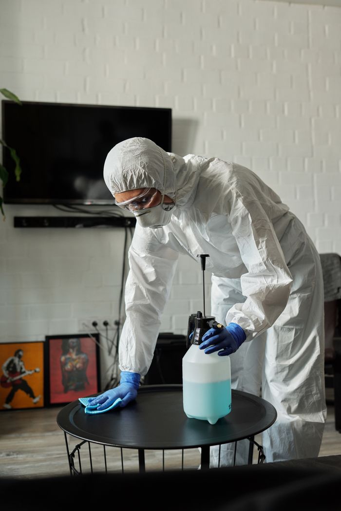 about-04 Person in protective gear cleaning a living room table with disinfectant.