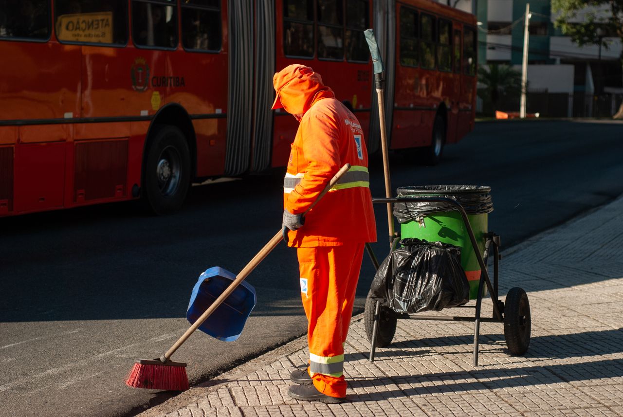 hero-img Street cleaner in bright orange uniform sweeping sidewalk near public bus in city setting.